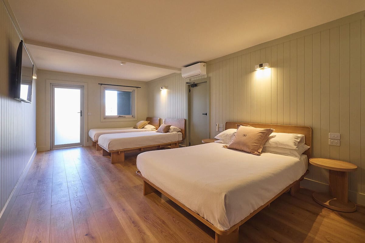 Habitación de hotel con dos camas individuales, una cama grande, suelo de madera, una ventana y un equipo de aire acondicionado.