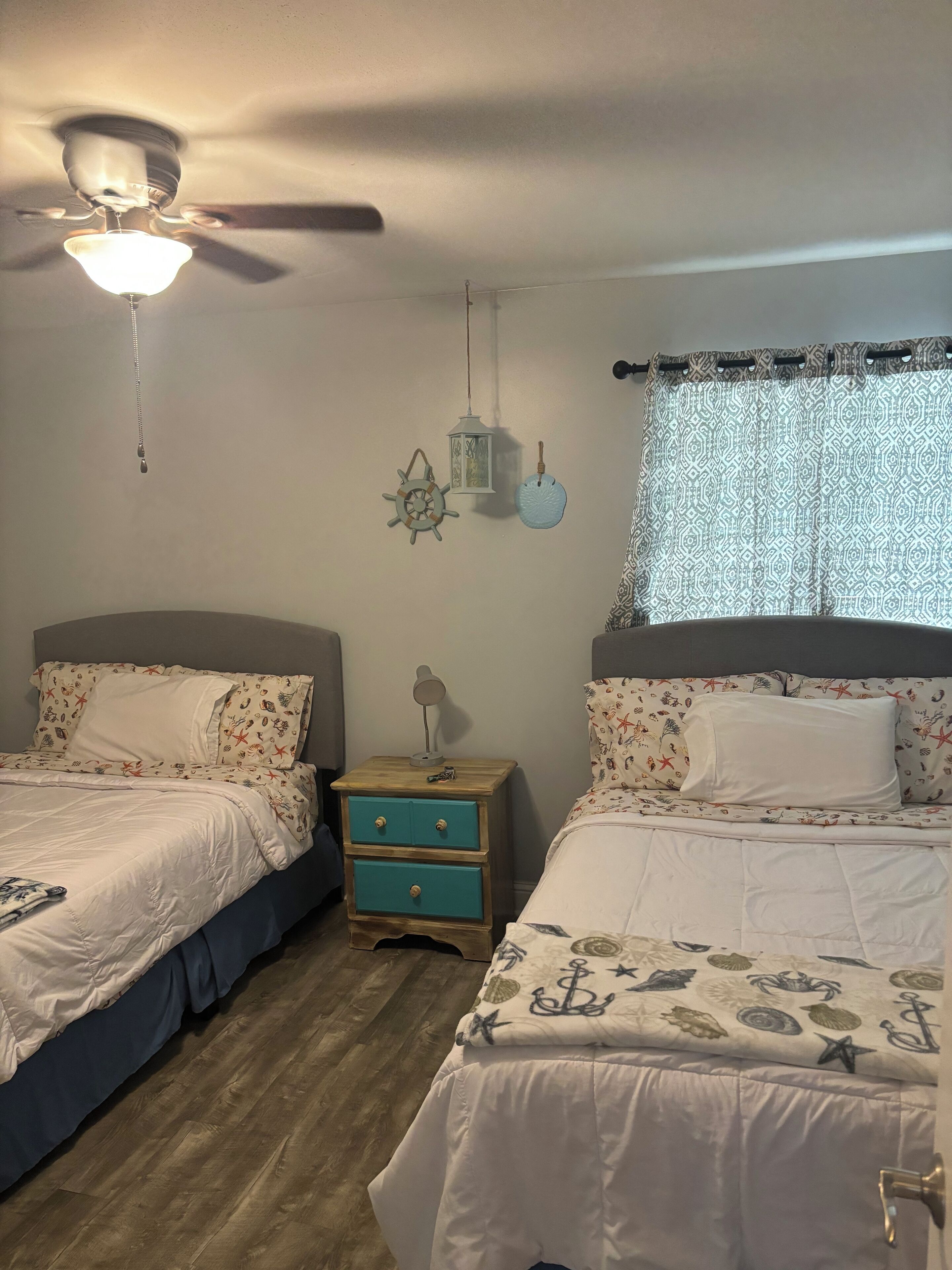 A bedroom with two beds, a ceiling fan, a nightstand with a lamp, and a window with patterned curtains.