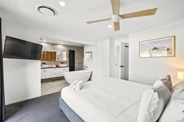 Une chambre moderne avec un grand lit, un ventilateur de plafond, un téléviseur à écran plat, une salle de bain avec baignoire et un coin cuisine visible à travers une porte ouverte.