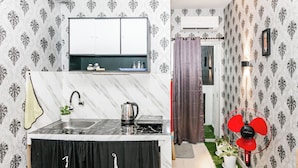 A compact kitchen with a marble countertop, a sink, and a kettle. There is a small window with a curtain, and a red fan is visible in the corner. - Hotel O Mp Room (Depok)