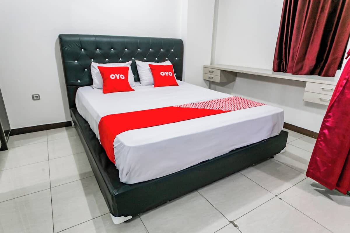 A neatly made bed with a red and white bedspread and matching pillows, a dark green upholstered headboard, and a white bedside table with a drawer.