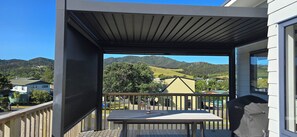 A balcony with a table and chairs, overlooking a scenic landscape with hills and houses. - The Beach Hut - A Walk to Hihi Beach. (Hihi)