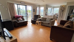 Ett rymligt vardagsrum med en stor soffa, en mindre soffa, ett soffbord och en hundsäng. I taket hänger en takfläkt, det finns ett tv-appar och ett köksområde som syns genom en öppen dörr.