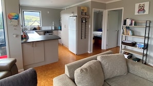 Ett modernt vardagsrum med en grå soffa, en svart fåtölj och ett soffbord. I bakgrunden syns ett kök med vita skåp och en vinhylla.