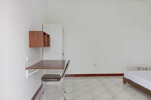 A small, sparsely furnished room with a wooden desk, a chair, a bed, and a small table. - OYO 94070 The Cozzy Exclusive Syariah (Bandar Lampung)