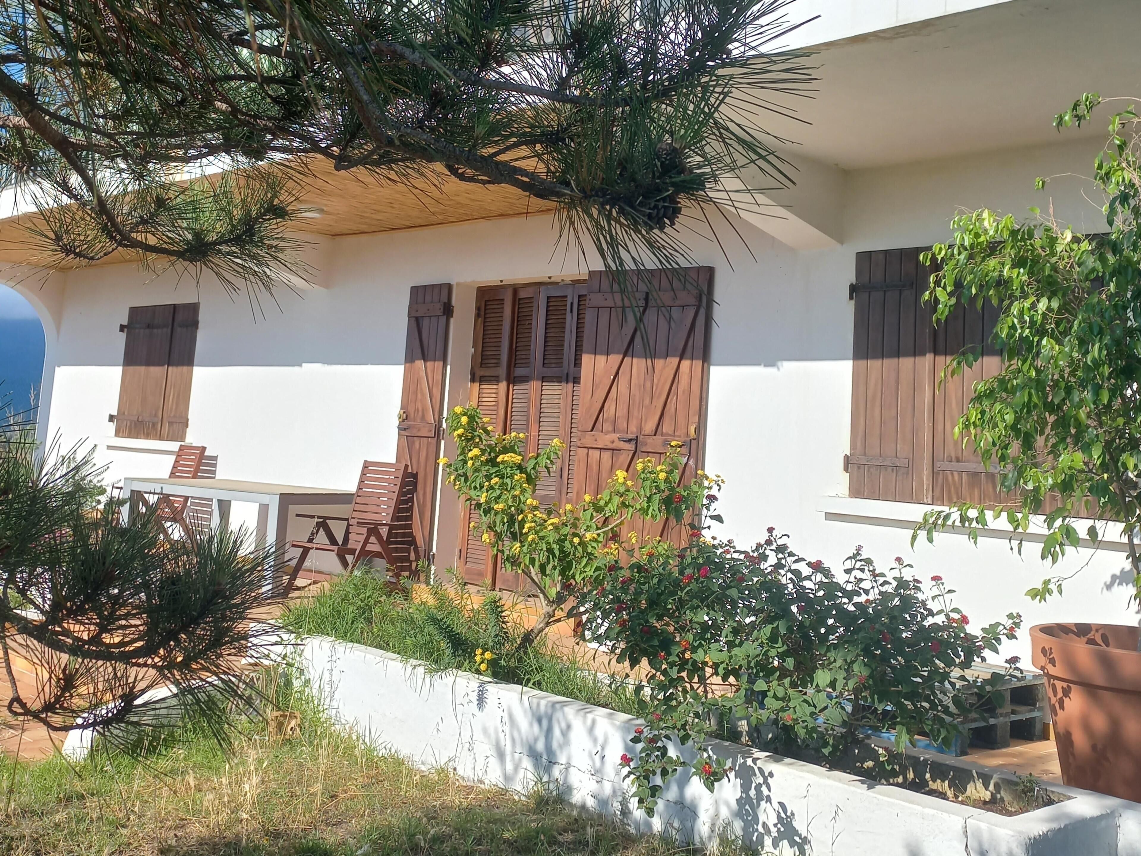 Un bâtiment blanc avec des volets en bois, un bac à fleurs en béton orné de fleurs jaunes et une table en bois avec des chaises.