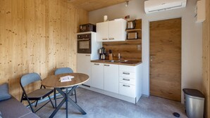 Una cucina compatta con mobili bianchi, un forno a microonde, una macchina da caffè e un lavandino. Si nota una porta in legno e un cestino per i rifiuti.