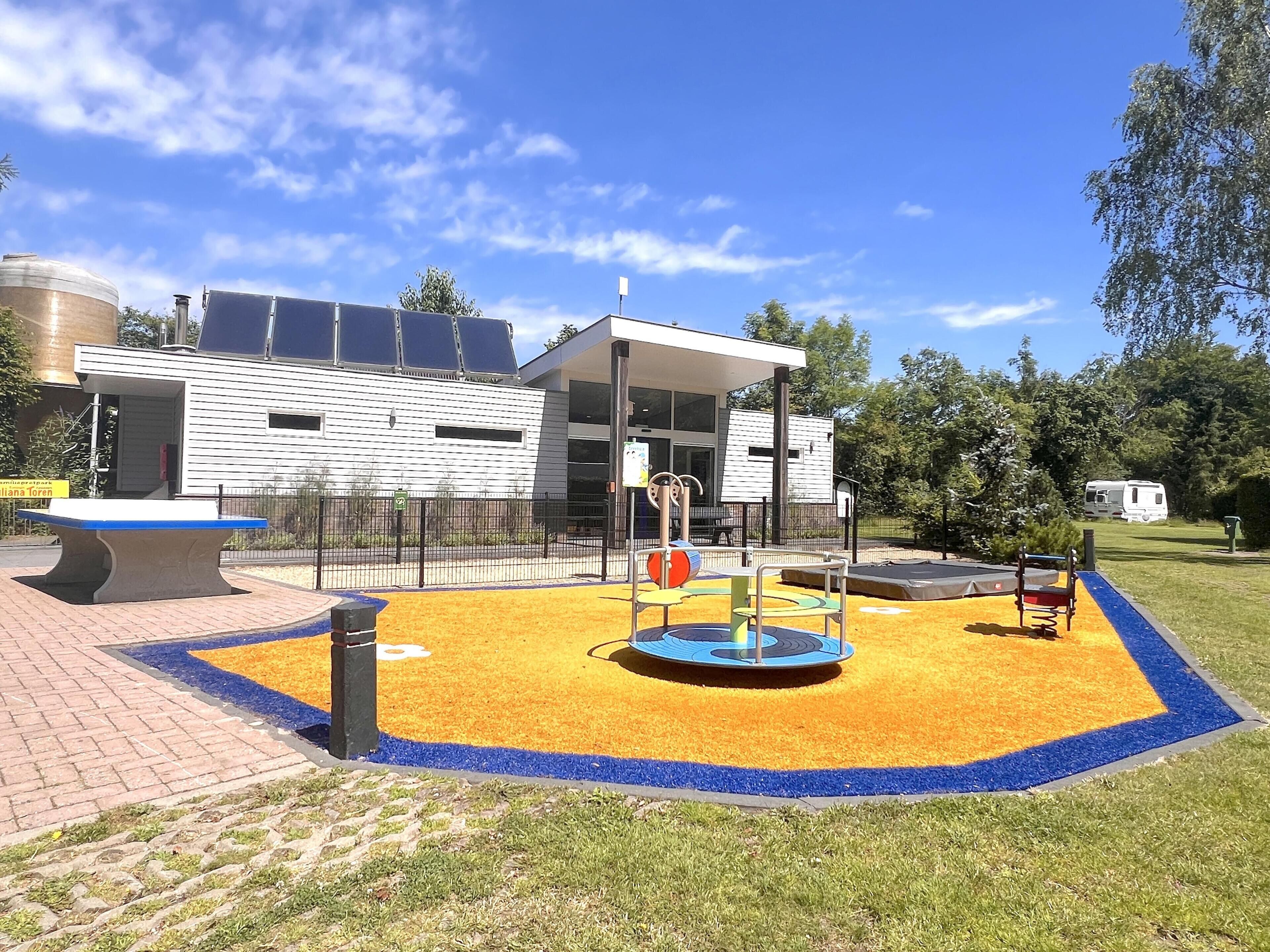 Une aire de jeux avec une zone de jeux aux couleurs bleu et jaune, un banc et un bâtiment doté de panneaux solaires en arrière-plan.