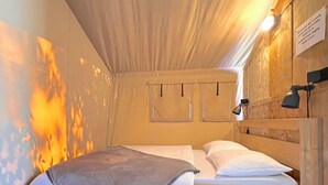 Un lit simple avec une couverture grise, une tête de lit en bois et une lampe de chevet, dans une pièce aux murs en bois et au plafond en pente.