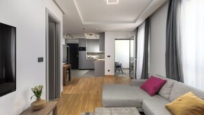 Un salon moderne avec un canapé gris, une table basse en bois et un téléviseur sur le côté gauche. On aperçoit une cuisine à l’arrière-plan, avec des meubles gris et un coin repas.