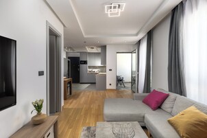 Una sala de estar moderna con un sofá gris, una mesa de centro de madera y un televisor a la izquierda. Se ve una cocina al fondo, con gabinetes grises y una zona de comedor.