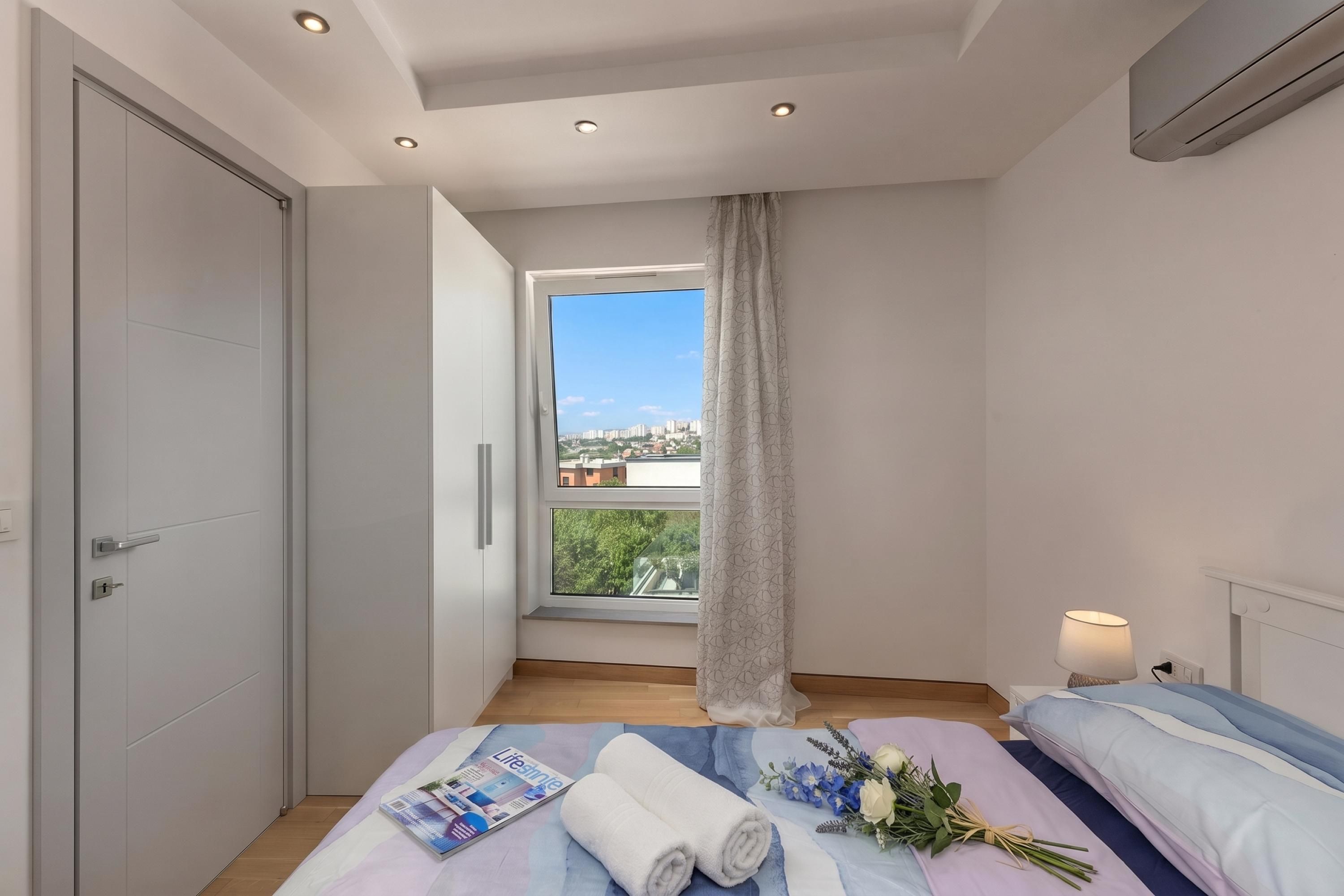 A modern bedroom with a large window, a bed with a magazine and towels, and a nightstand with a lamp.