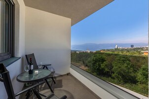A balcony with a table and chairs, a bottle of wine, and two glasses. The balcony overlooks a cityscape with buildings and trees.