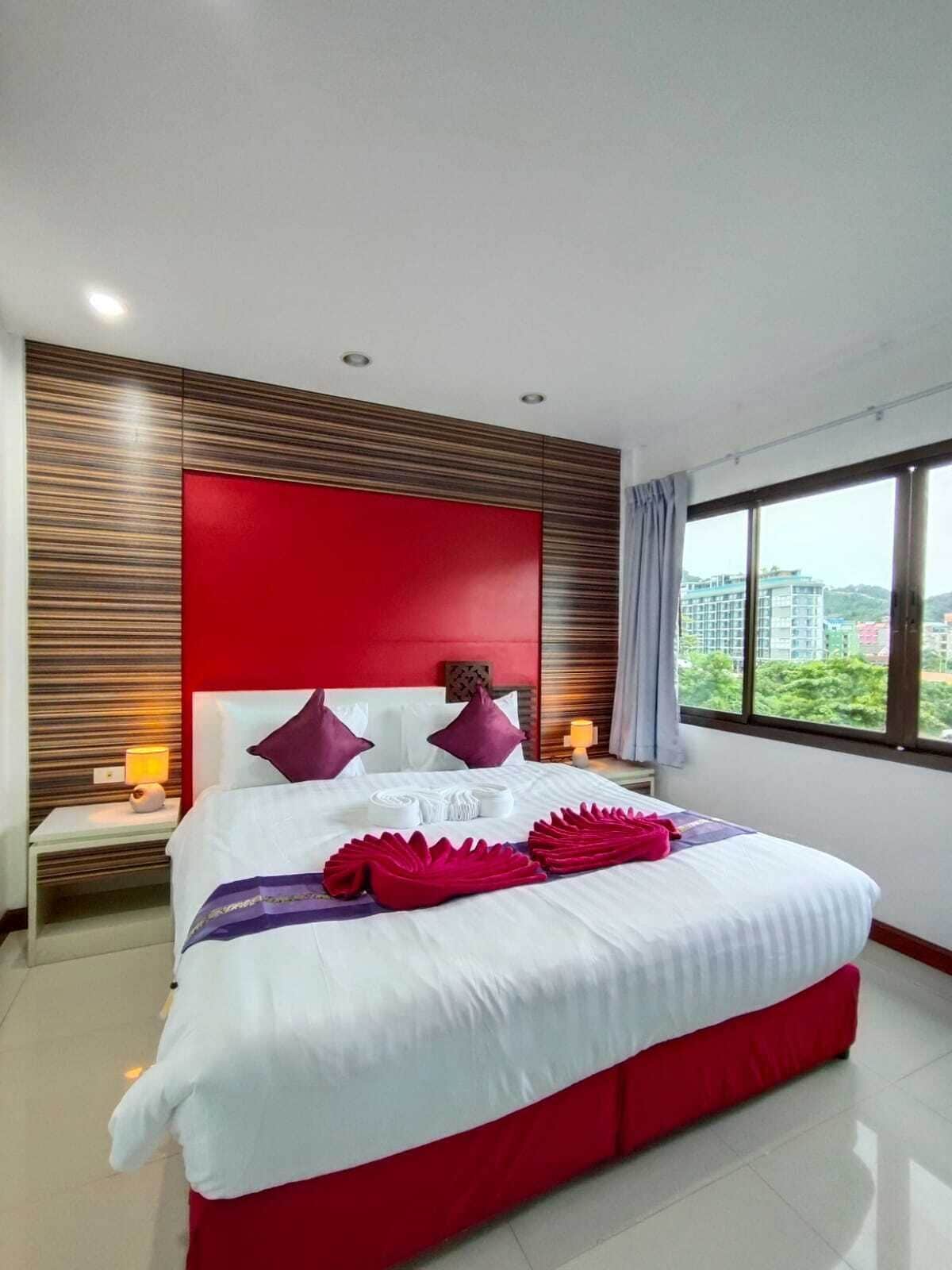 A modern hotel room with a large bed, red accents, and a city view through the window.