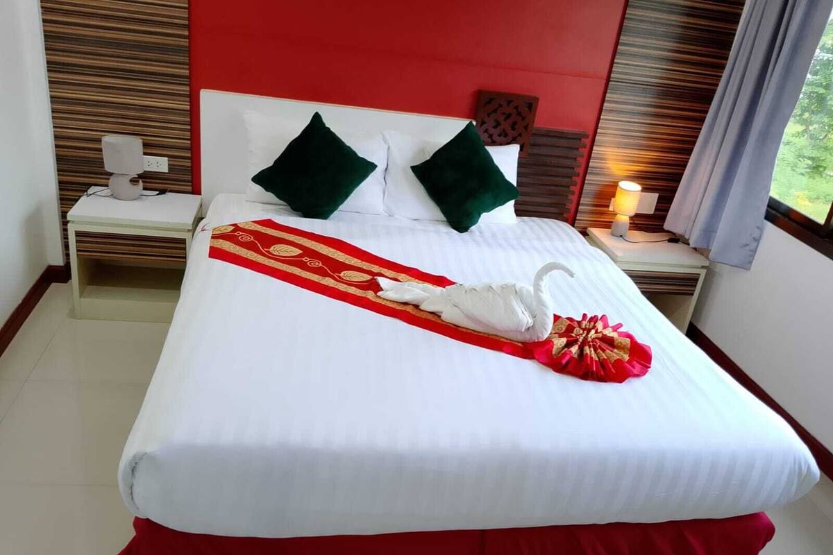 A hotel room with a neatly made bed, a red and white bedspread, two green pillows, a wooden headboard, and a nightstand with a lamp.