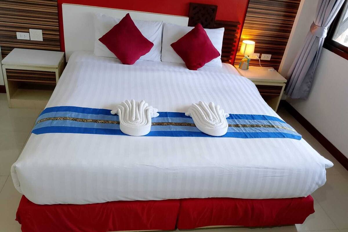 A hotel room with a large bed, two red pillows, a white headboard, a nightstand with a lamp, and a red skirted bed base.