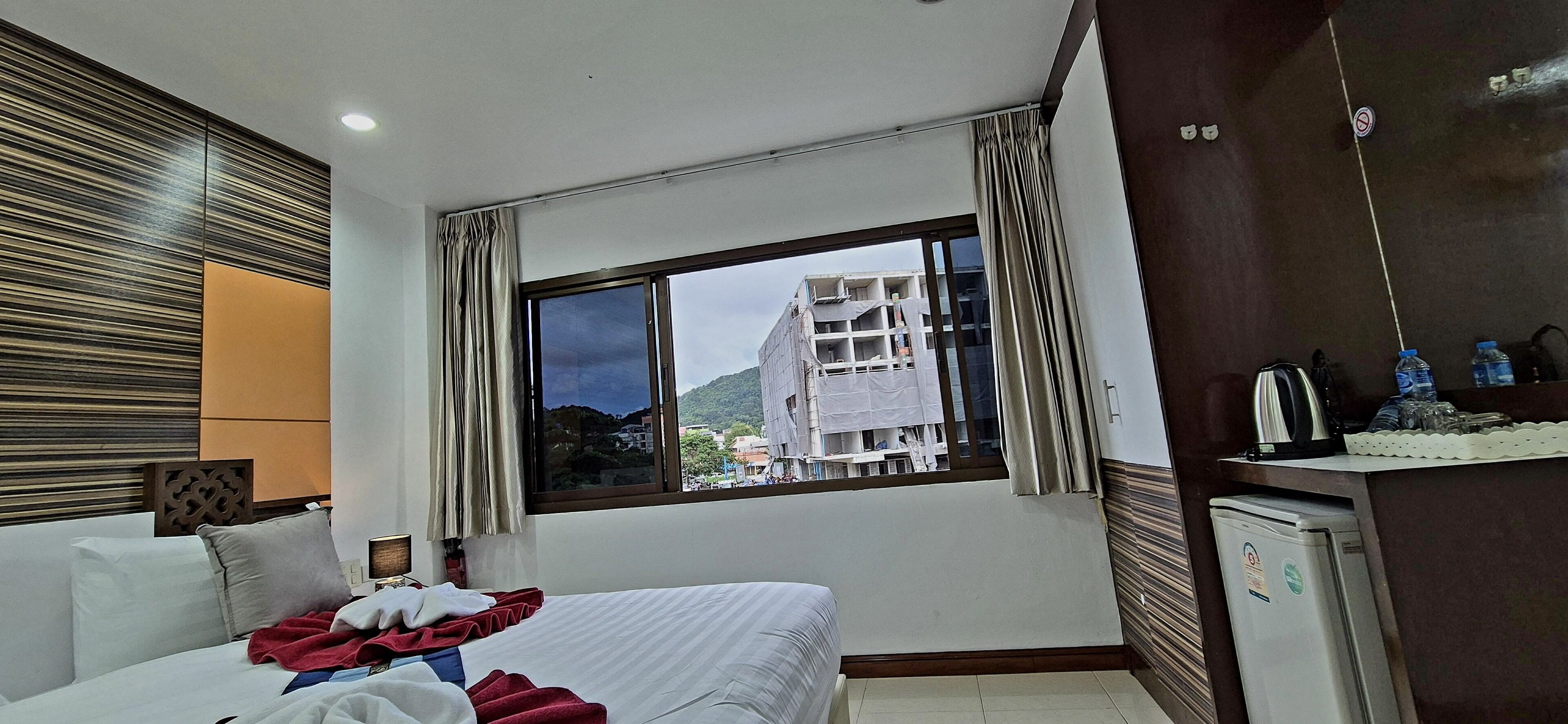 Habitación de hotel con cama, escritorio, refrigerador y ventana con vista a edificios y montañas.