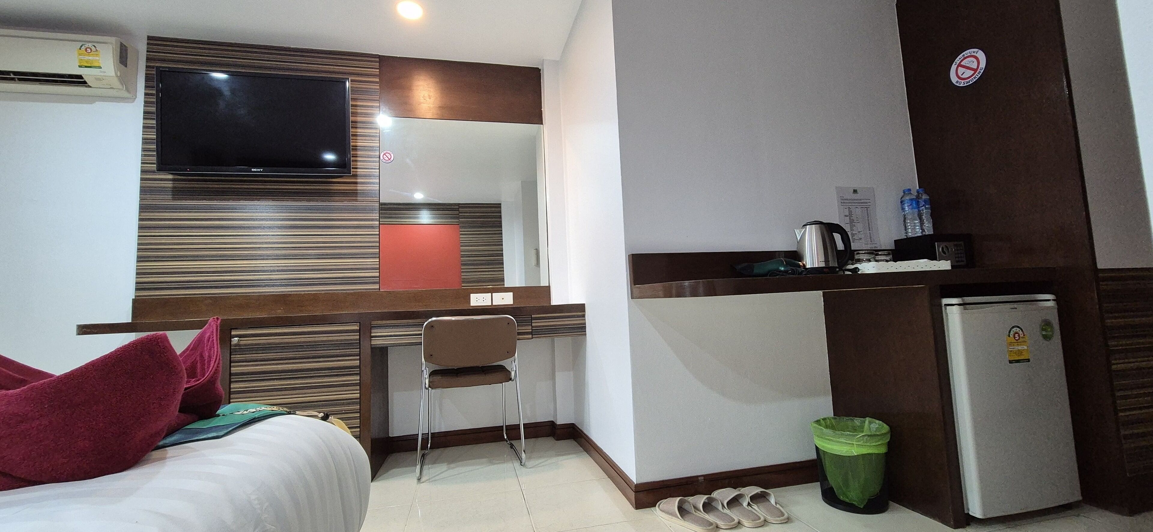A hotel room with a bed, a desk with a chair, a television, a small kitchenette, and a trash bin.