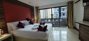A hotel room with a large bed, a desk, and a balcony with a view of buildings. - Patong Max Value Hotel (Patong)