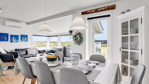 Ein moderner Essbereich mit einem Tisch für sechs Personen, umgeben von grauen Stühlen. Eine weiße Decke mit Holzbalken, ein großes Fenster mit Blick ins Grüne und eine weiße Schrank mit Glastüren sind zu sehen.