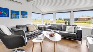 Ein modernes Wohnzimmer mit einem grauen Ledersofa, einem weißen Couchtisch und einem schwarzen Ledersessel. An der Wand hängen drei gerahmte Kunstwerke, und ein großes Fenster bietet einen Blick ins Freie.