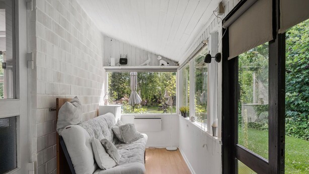 A cozy room with a white brick wall, a wooden floor, a white sofa with cushions, and a window overlooking a garden. - 4 bedroom amazing home in Martofte (Martofte)