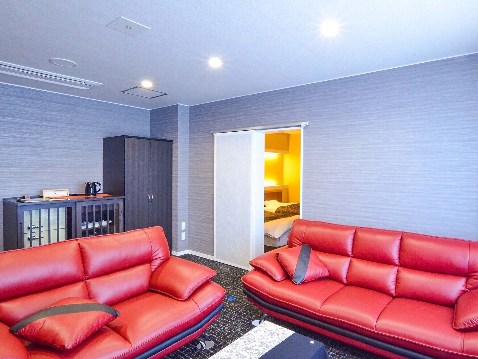 Une chambre d’hôtel avec deux canapés en cuir rouge, un buffet en bois foncé et un lit, le tout dans une pièce éclairée d’une lumière jaune à l’arrière-plan.