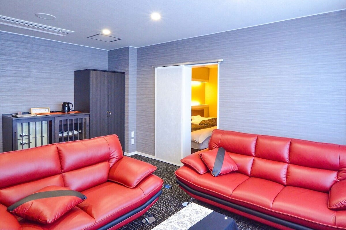 Une chambre d’hôtel avec deux canapés en cuir rouge, un buffet en bois foncé et un lit, le tout dans une pièce éclairée d’une lumière jaune à l’arrière-plan.