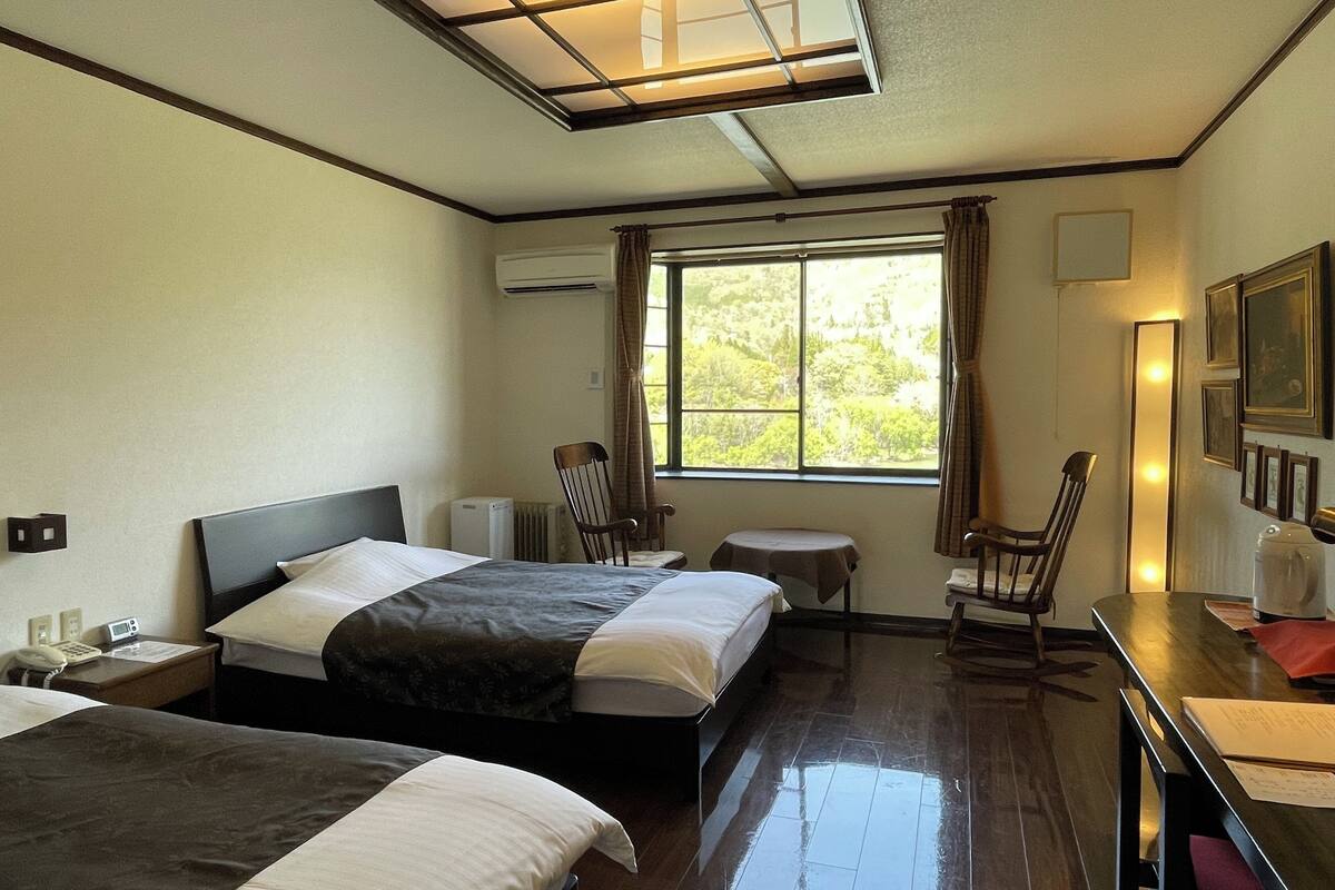 A hotel room with two beds, a desk, a chair, a small table, and a window with a view of trees.