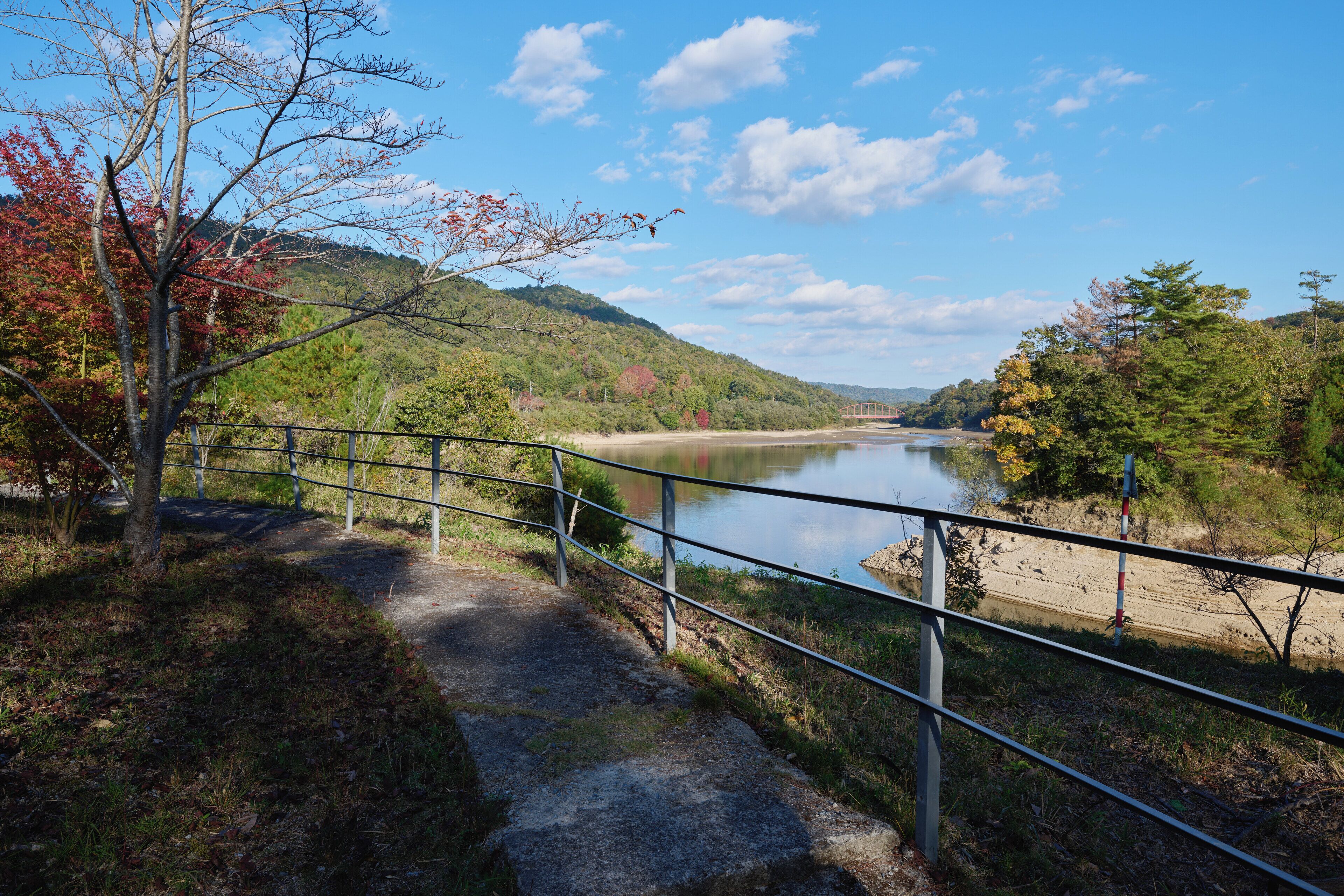 A pathway beside a river with a metal railing, surrounded by trees and hills.