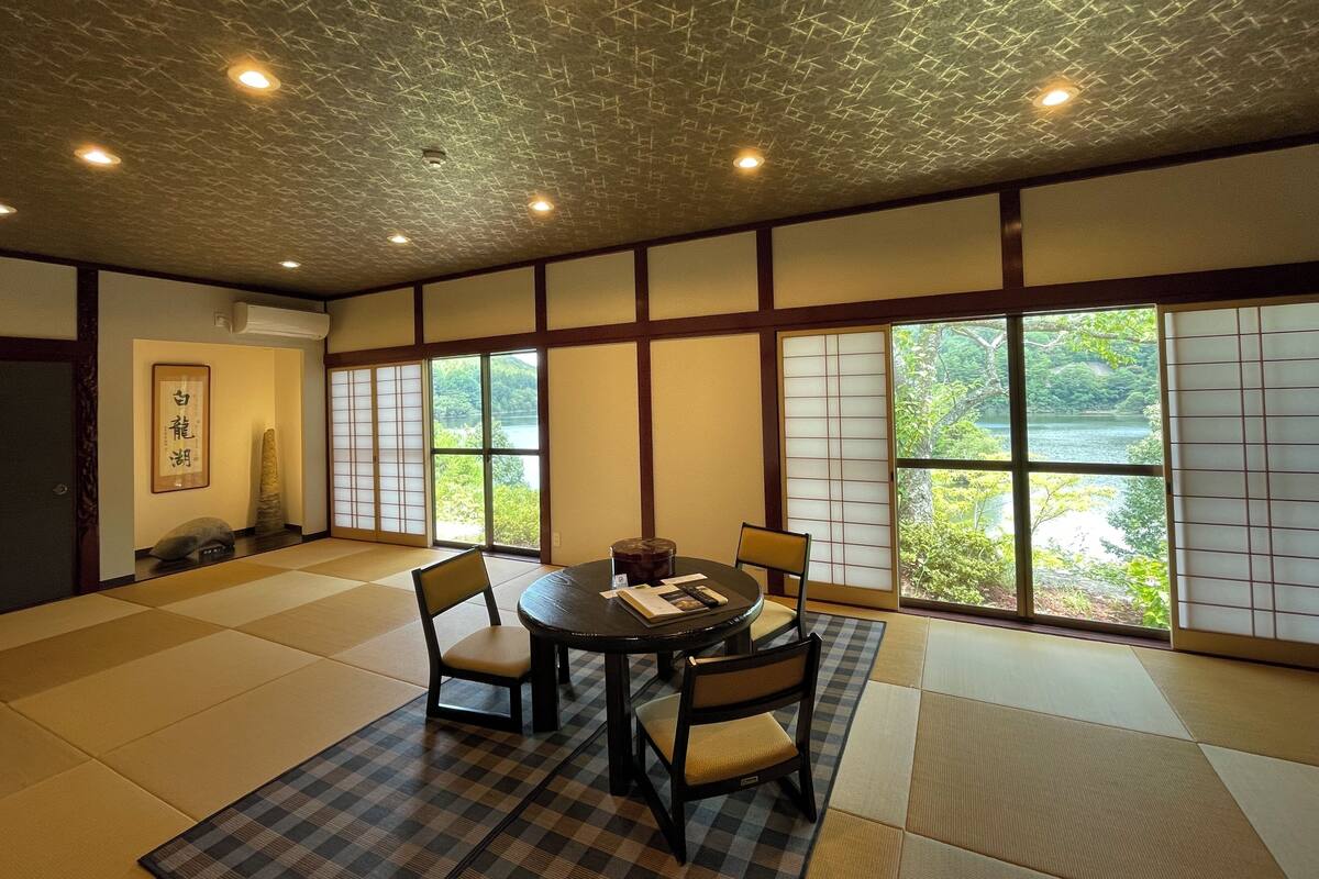 A traditional Japanese room with tatami flooring, a low table with chairs, and large windows with shoji screens.