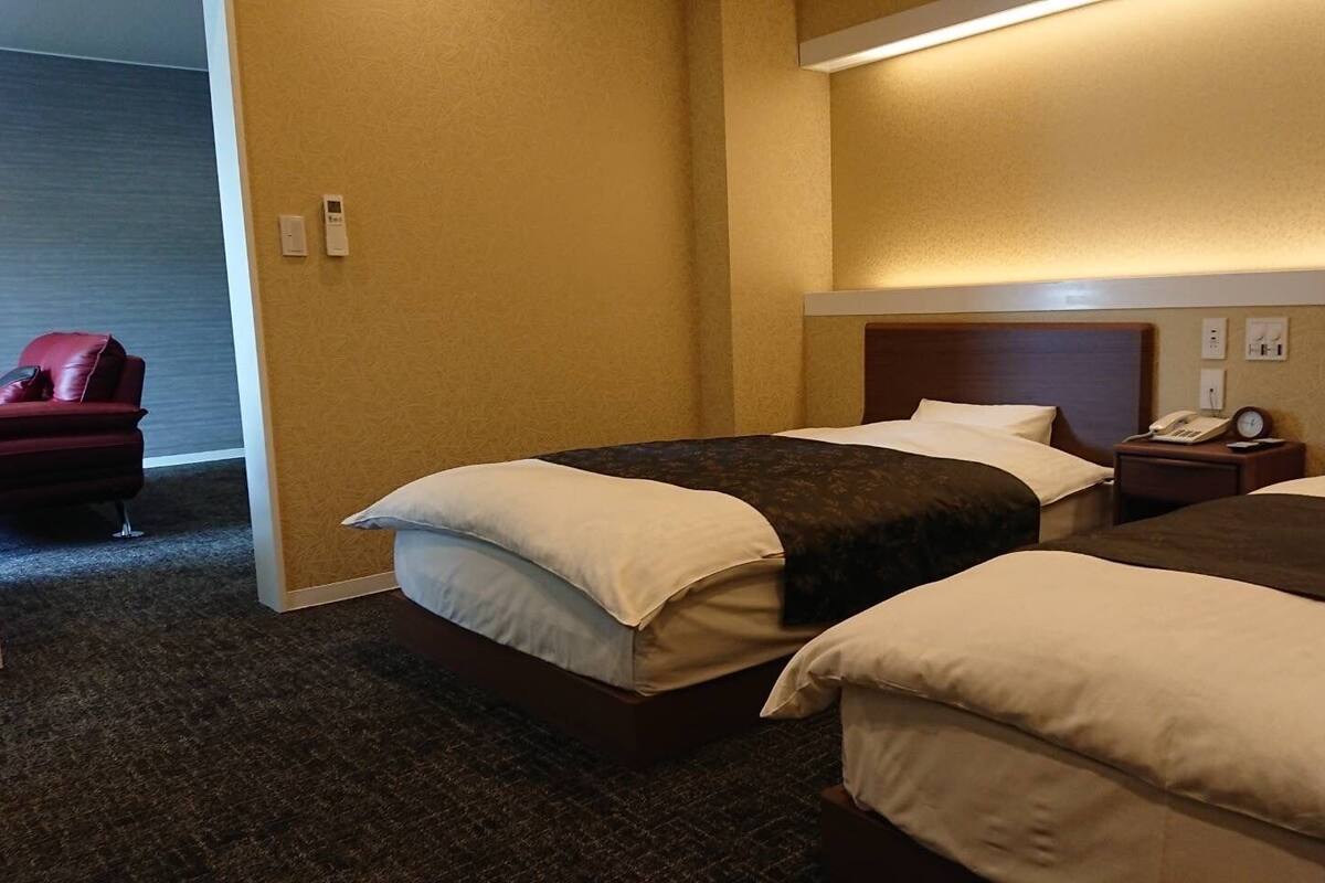 A hotel room with two beds, a red chair, a bedside table with a phone, and a wall-mounted light.