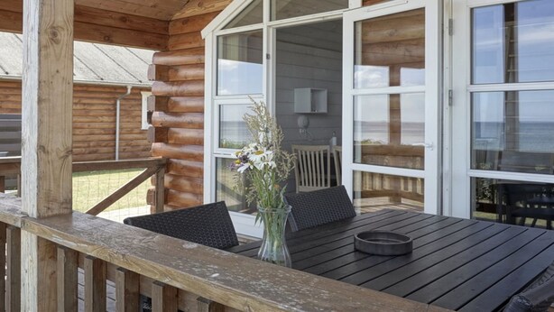 Een houten terras met een tafel en stoelen, grote ramen en uitzicht op de omgeving.