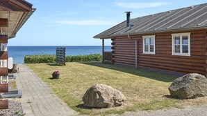 Een houten hutje met uitzicht op zee, een verhard pad en grote rotsen op de voorgrond.