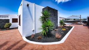 A modern building with white walls and a stone facade, surrounded by a landscaped garden with various plants and a pool area. - Lanzarote Paradise - Pool, AACC and BBQ By LVH (Playa Blanca)