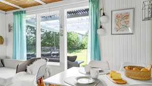 Une pièce lumineuse et bien éclairée, avec un plafond en bois, des murs blancs et une grande fenêtre ornée de rideaux turquoise. Une table à manger blanche, avec de l’assiette et un panier de pain, est dressée.