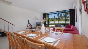 Un coin repas avec une table en bois dressée pour quatre personnes, entourée de chaises en bois. Un canapé rouge et un téléviseur sont visibles au fond.