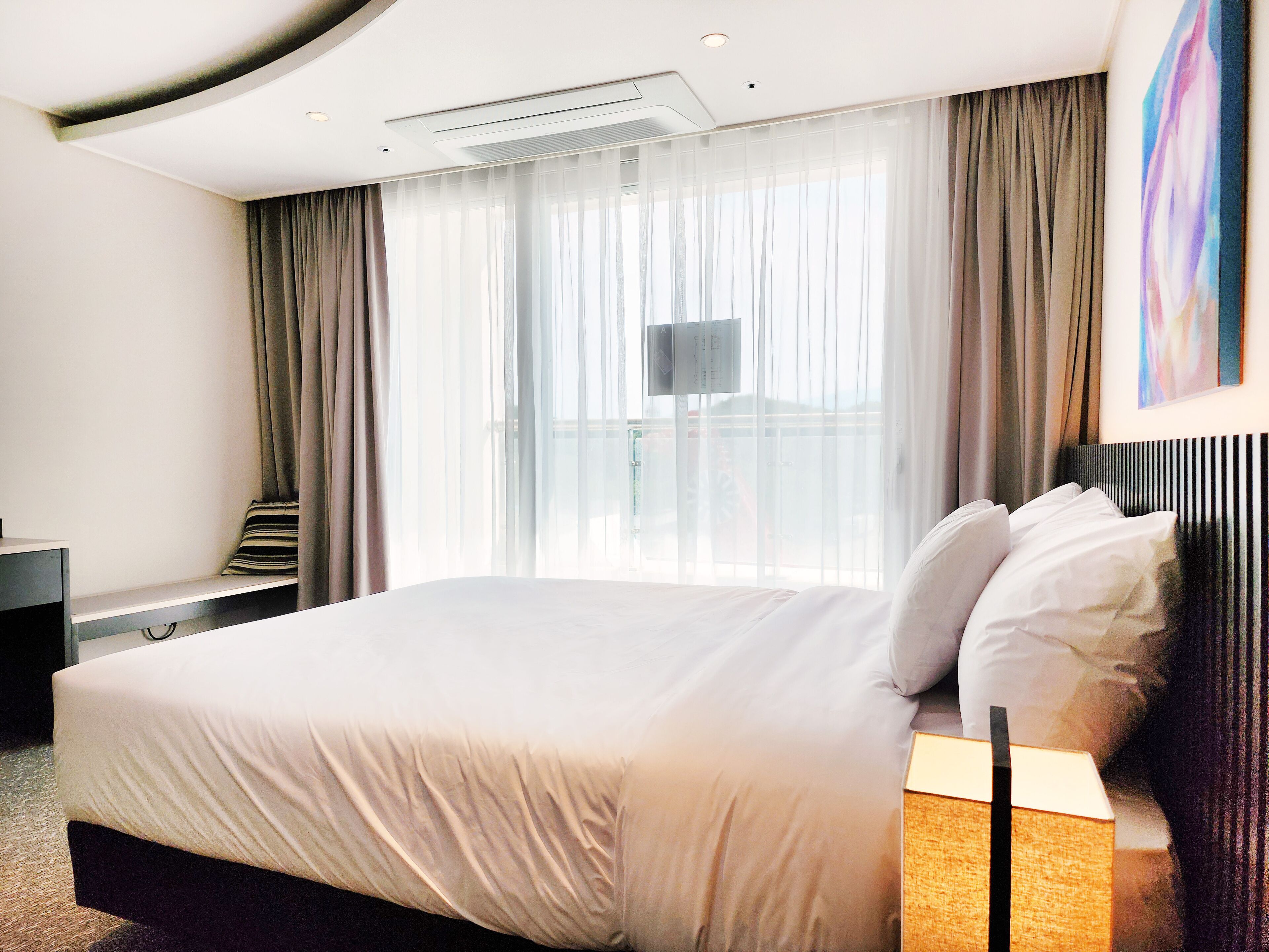 A modern hotel room with a large bed, a bedside table, a TV, and a view of the city through sheer curtains.