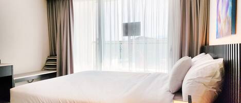 A modern hotel room with a large bed, a bedside table, a TV, and a view of the city through sheer curtains.