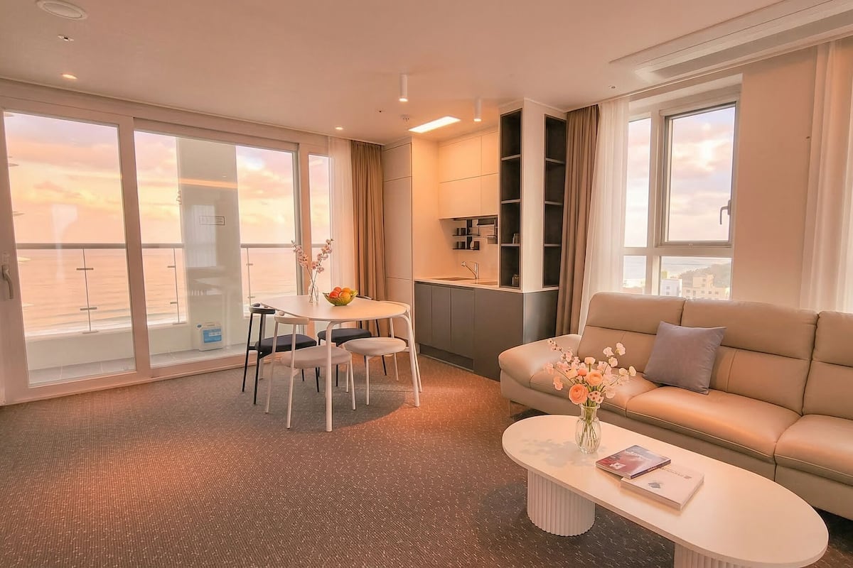 Une chambre d’hôtel avec un canapé, une table à manger avec des chaises, un petit coin cuisine et de grandes fenêtres.