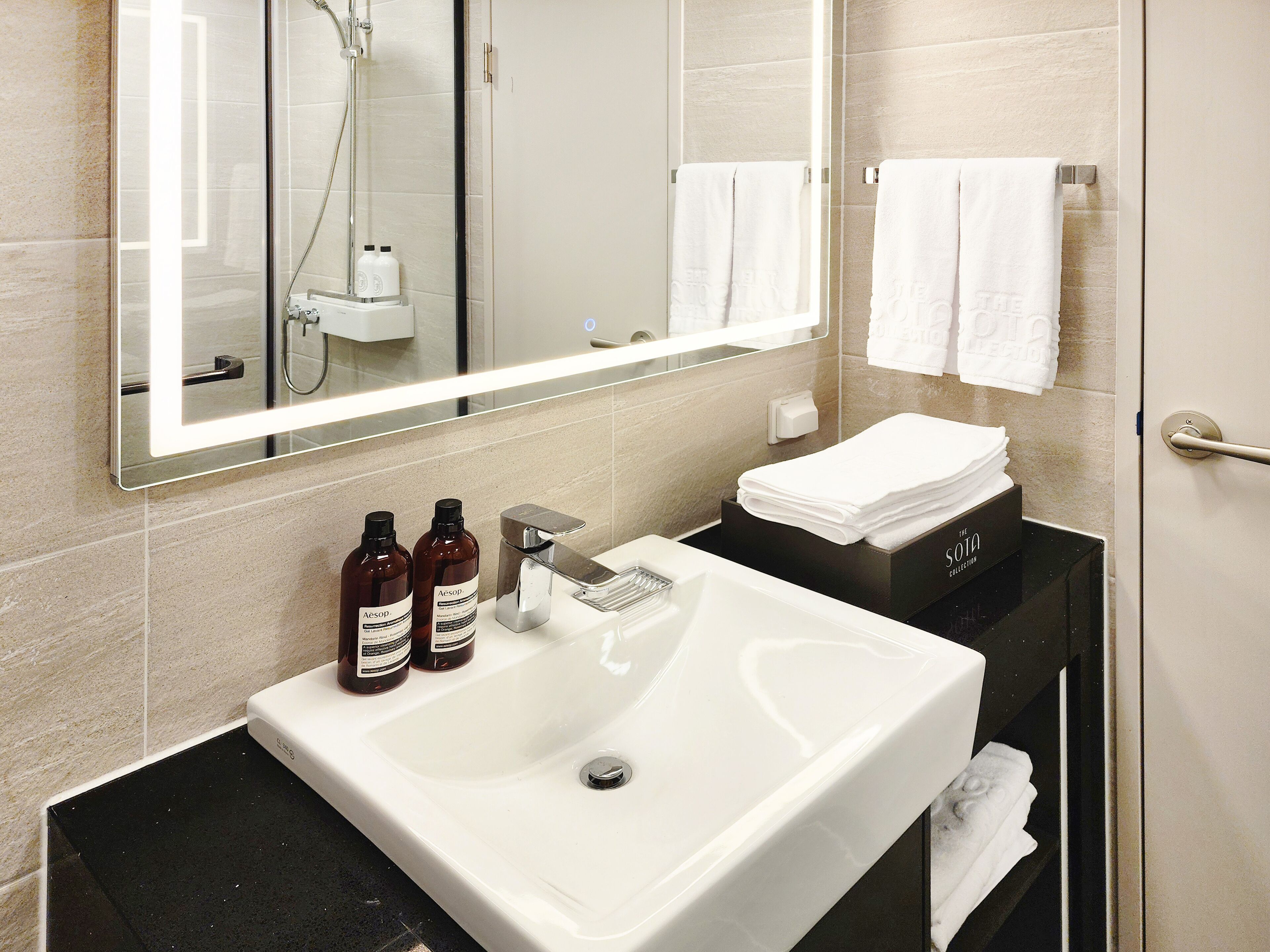 Et moderne badeværelse med et stort spejl, en hvid vask og to flasker Aesop shampoo.