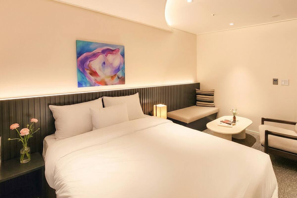 Une chambre d’hôtel avec un grand lit, une table de chevet avec une lampe, une chaise, une petite table avec un vase et un tableau abstrait aux couleurs vives accroché au mur.