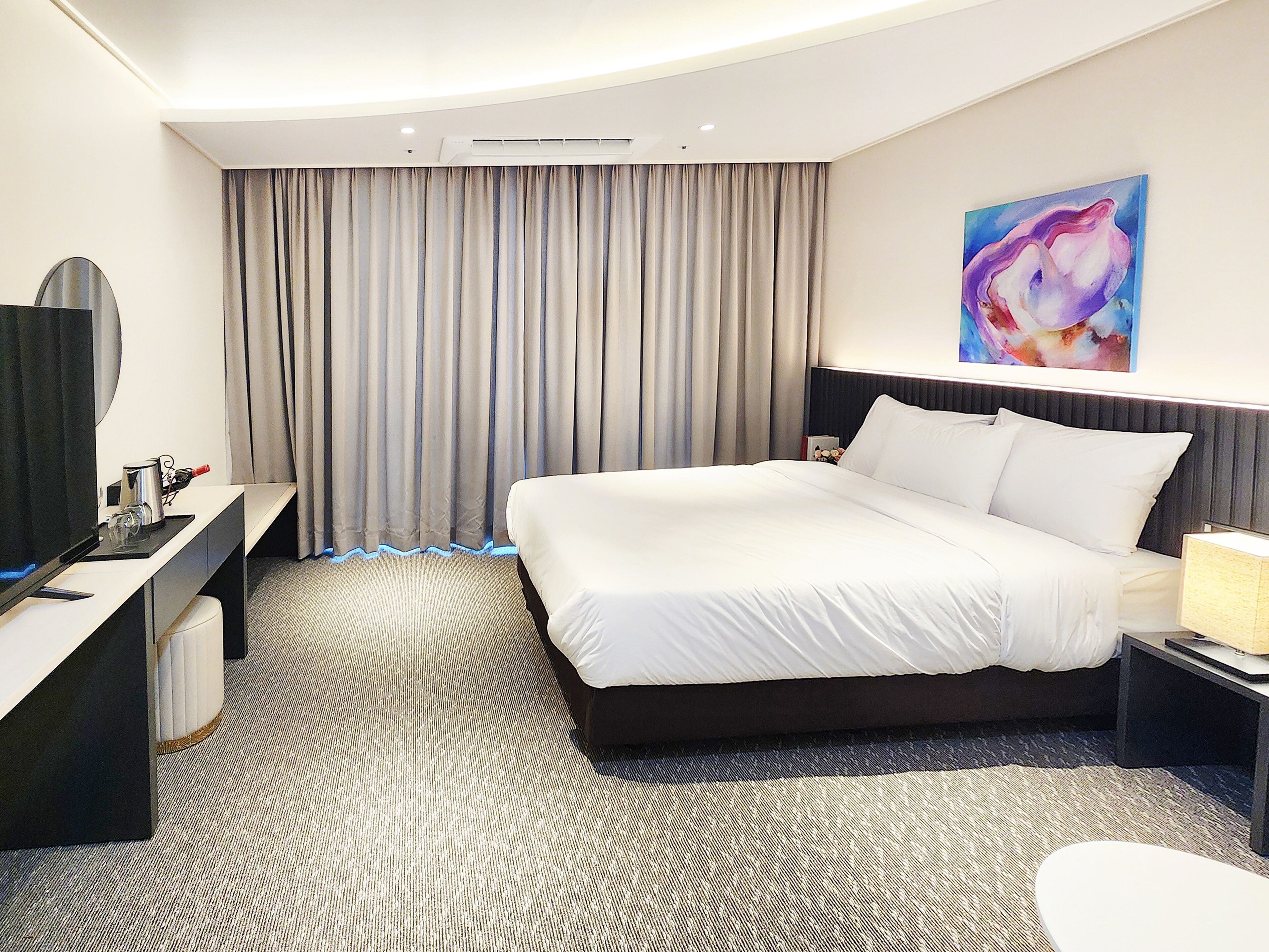 A hotel room with a bed, a desk, a chair, a TV, and a painting on the wall.