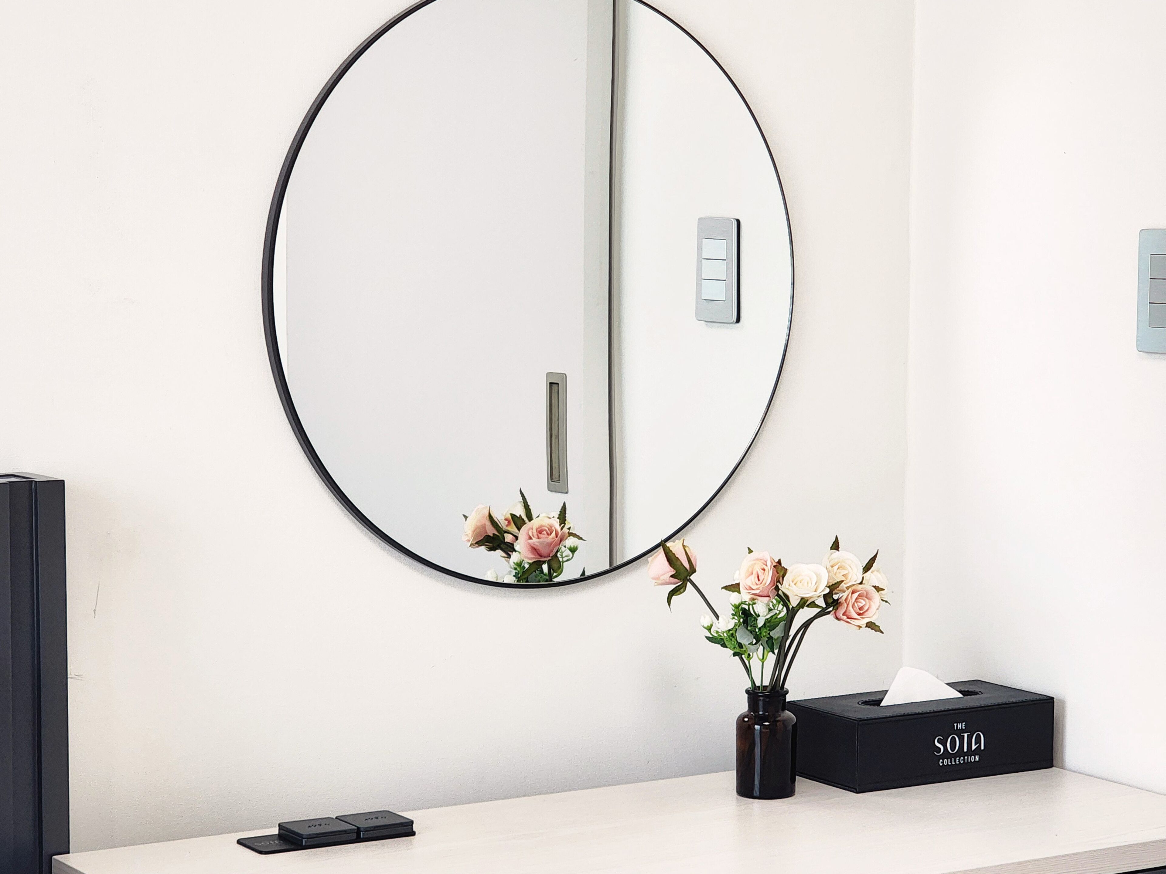 Un baño con un espejo redondo, un florero negro con flores y una caja de pañuelos en la encimera con la etiqueta 'THE SOTA COLLECTION'.