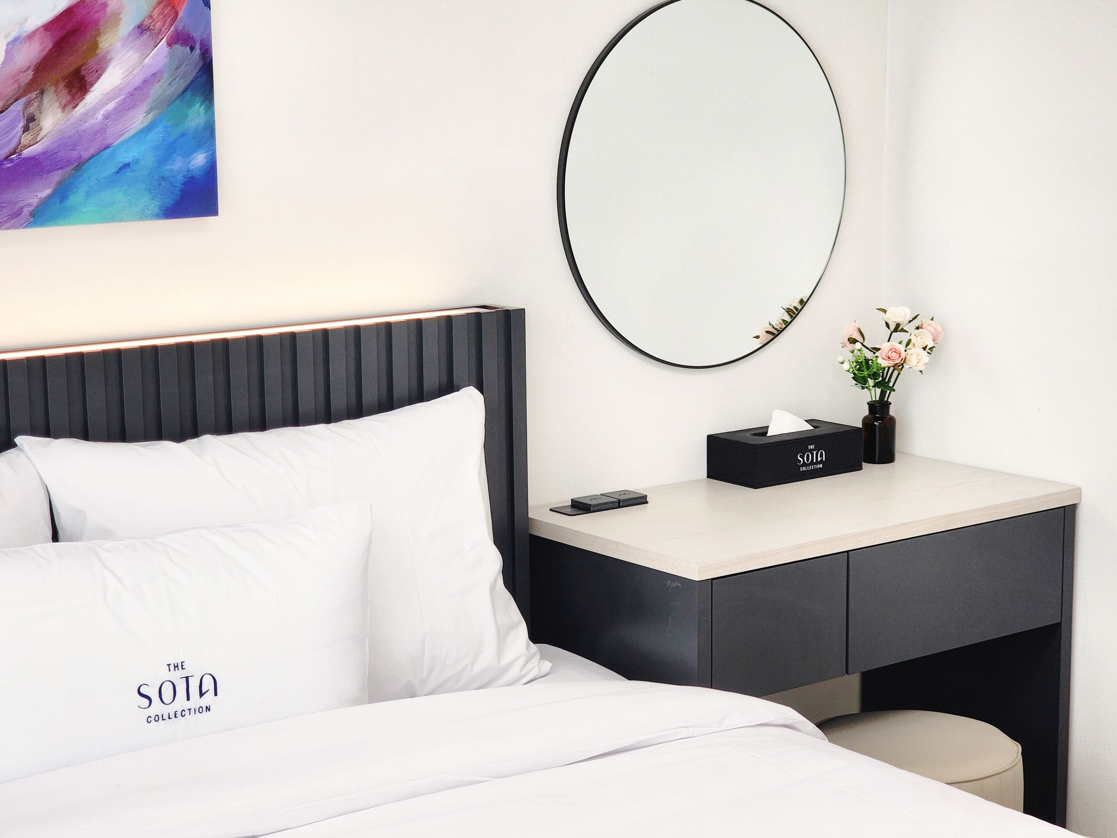 A modern hotel room with a neatly made bed, a bedside table with a tissue box and a vase of flowers, and a large mirror on the wall.