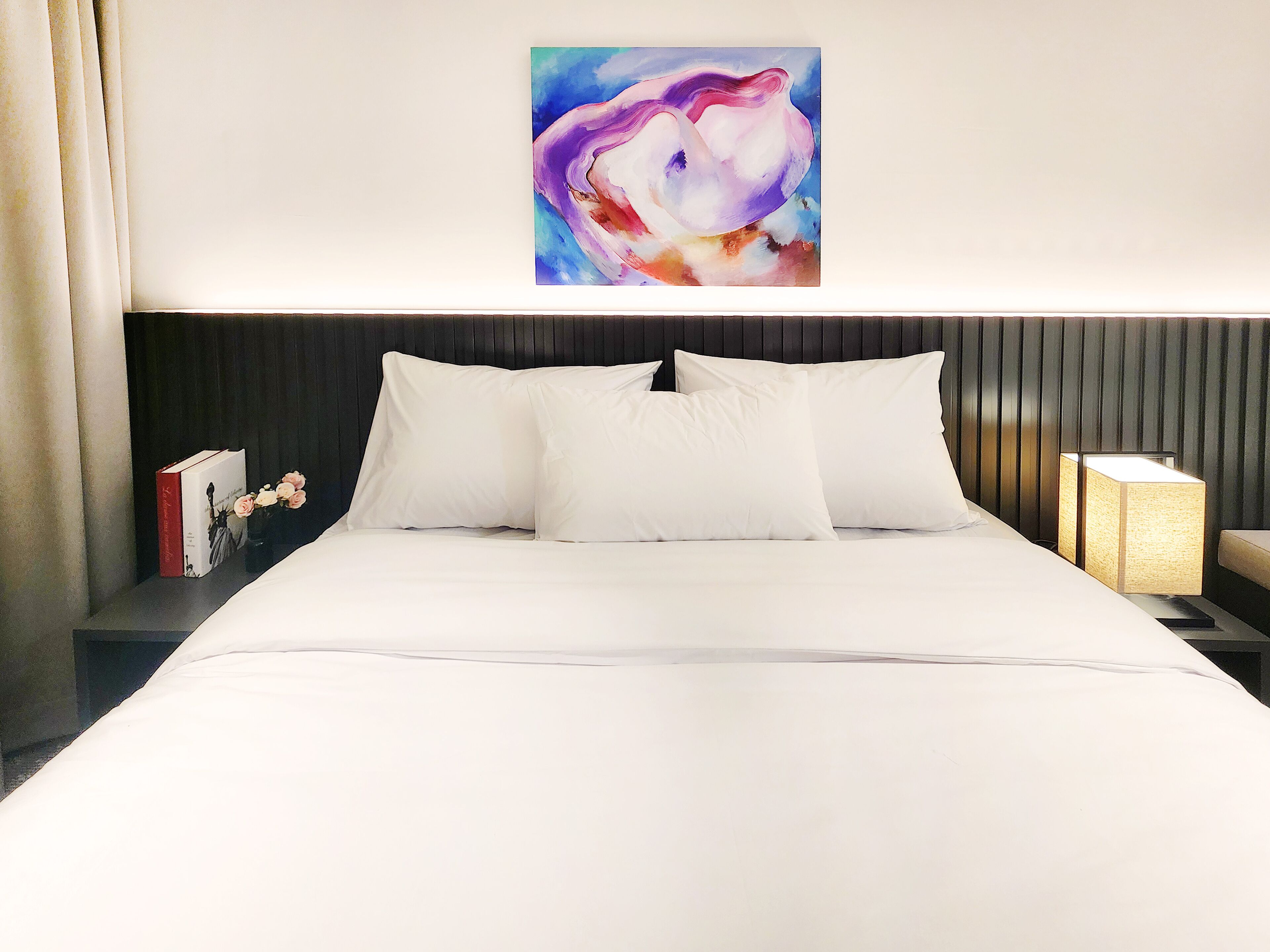 Een net opgemaakt bed met witte lakens, een donker hoofdbord en een schilderij erboven.