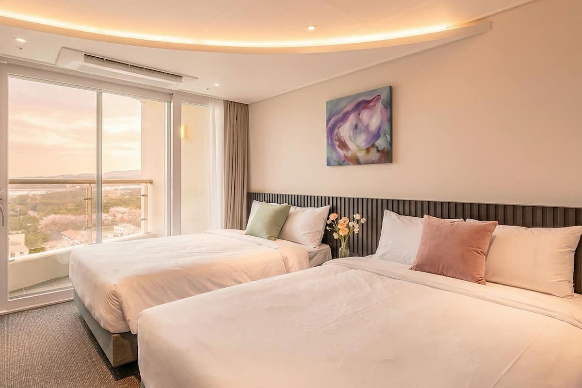 Une chambre d’hôtel avec deux lits, une grande fenêtre donnant sur un beau paysage, un tableau au mur et un vase rempli de fleurs.