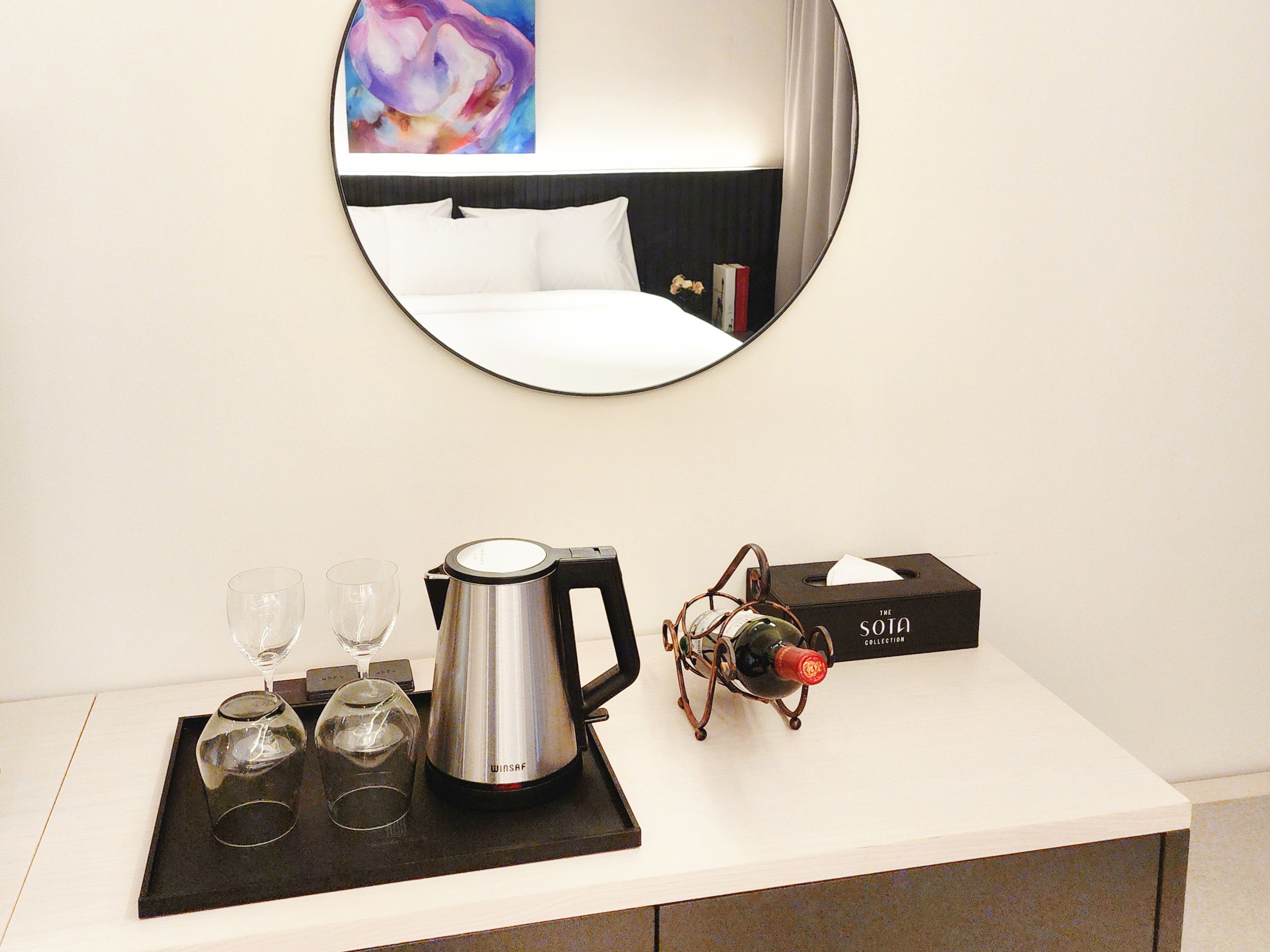 A hotel room with a bed, a bedside table with a coffee maker, wine glasses, and a box labeled "The Sota Collection".