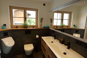 Une salle de bain moderne avec un grand miroir, des toilettes blanches, un lavabo avec un robinet noir et deux plantes en pot sur le rebord de la fenêtre.