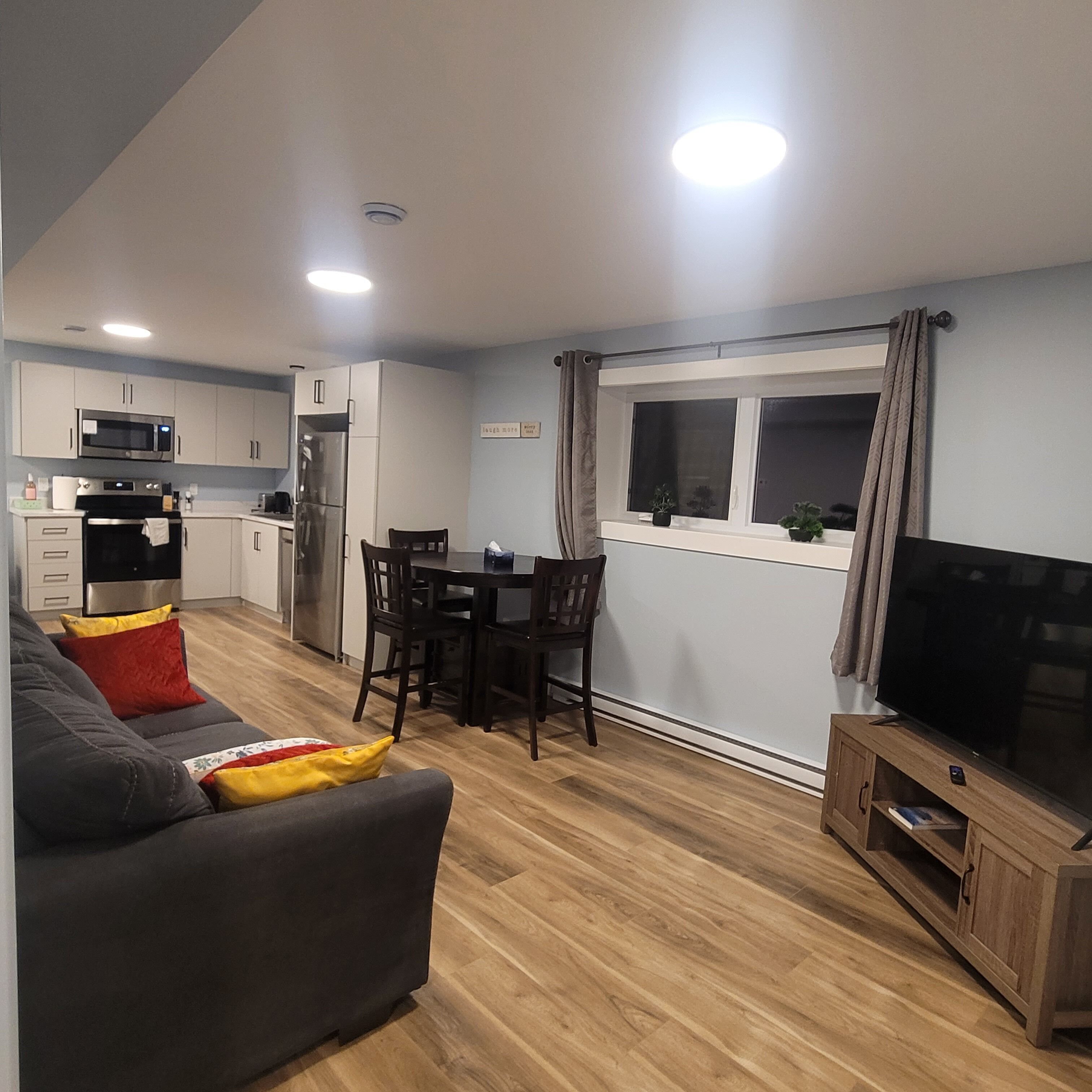Una cocina moderna con armarios blancos, microondas integrado y refrigerador de acero inoxidable. Cuenta con un comedor con mesa y sillas, y un televisor sobre un mueble de madera. El espacio tiene piso de madera y un sofá gris con cojines rojos y amarillos.